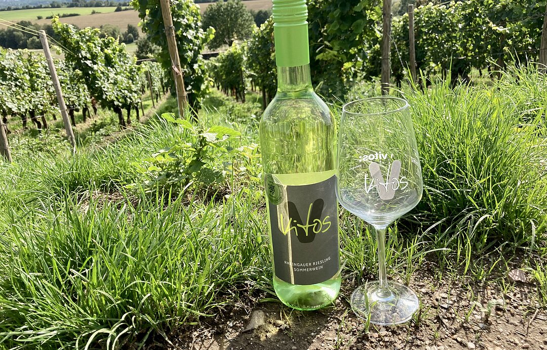 Ein Weinglas und eine Weinflasche stehen auf einer Wiese, dahinter Weinberge